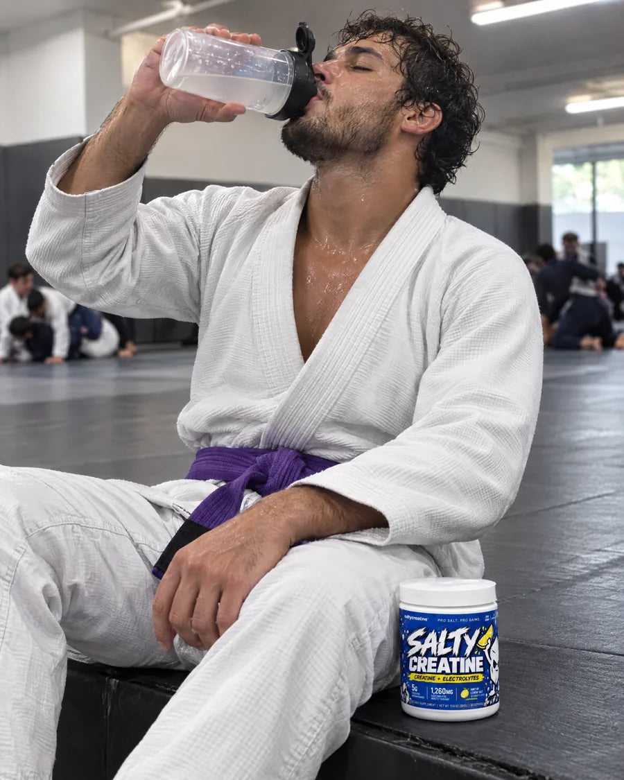 A jiu-jitsu athlete in a sweaty gi drinking from a shaker next to a tub of Salty Creatine
